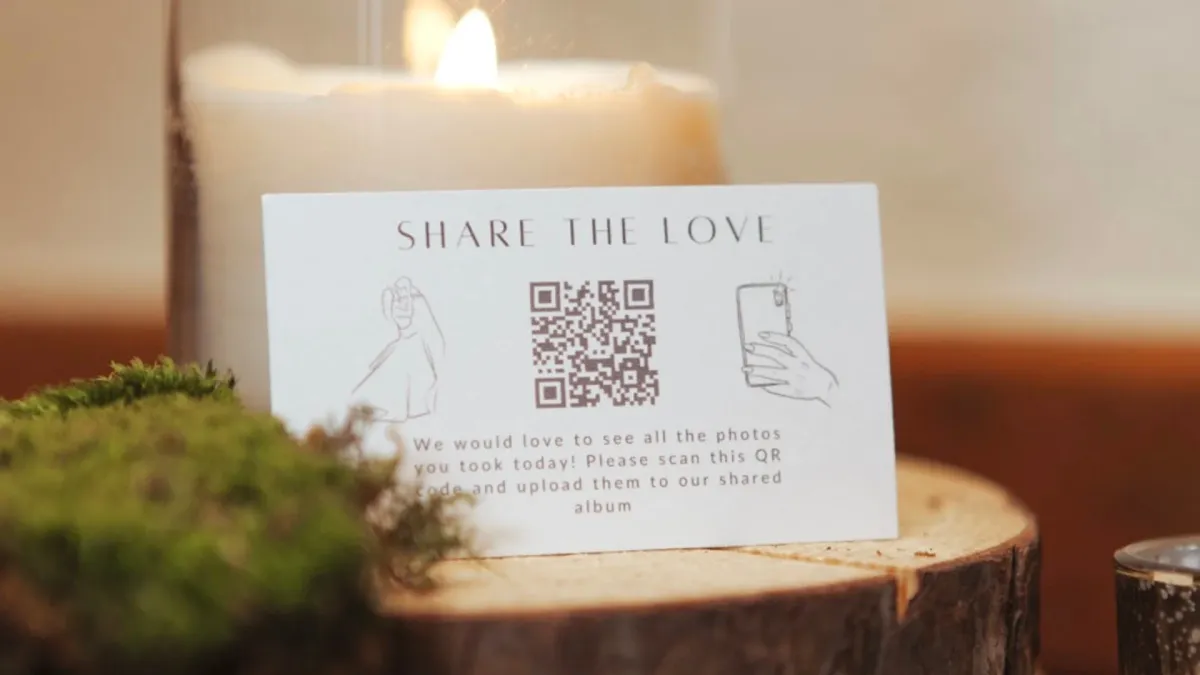 A printed QR code card on a wooden table at a wedding reception, asking guests to scan and share their photos